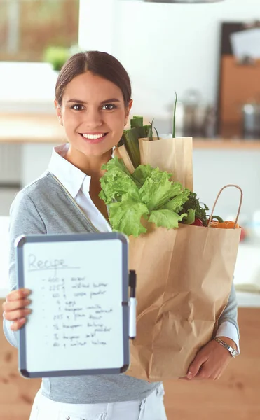 Mujer joven sosteniendo bolsa de la compra de comestibles con verduras. De pie en la cocina — Foto de Stock