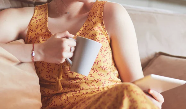 Νεαρή γυναίκα χρησιμοποιεί tablet ενώ χαλαρώνει στο σπίτι με καφέ στον καναπέ καναπέ — Φωτογραφία Αρχείου