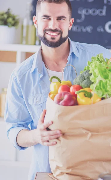 Egy férfi, aki papírzacskót tart tele élelmiszerekkel a konyhai háttérben. Vásárlás és egészséges étel koncepció — Stock Fotó