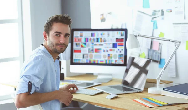 Retrato del joven diseñador sentado en el estudio gráfico frente a la computadora portátil y el ordenador mientras trabaja en línea. — Foto de Stock