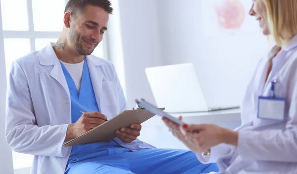 Médico bonito está conversando com jovem médica e fazendo anotações enquanto está sentado em seu escritório. — Fotografia de Stock