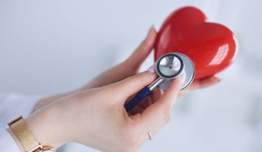 Steteskoplu bir doktor, beyaz arka planda kırmızı kalbi inceliyor..
