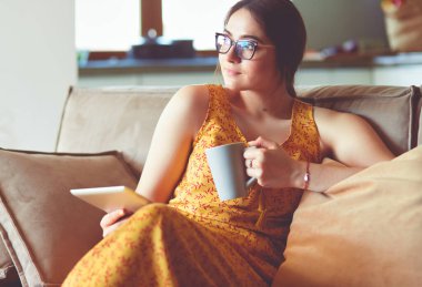 Genç kadın evinde kanepede kahve içerken tablet kullanıyor..