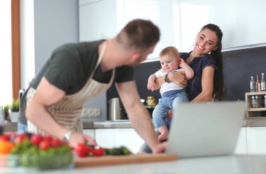 Masadaki dizüstü bilgisayara bakarken mutfakta sebze pişiren adamın portresi..