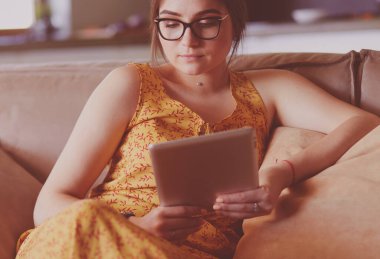 Genç kadın evinde kanepede kahve içerken tablet kullanıyor..