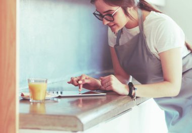 Mutfaktaki genç, beyaz tenli esmer ev kadınının portresi. Sabah bir fincan kahve ve tabletle.