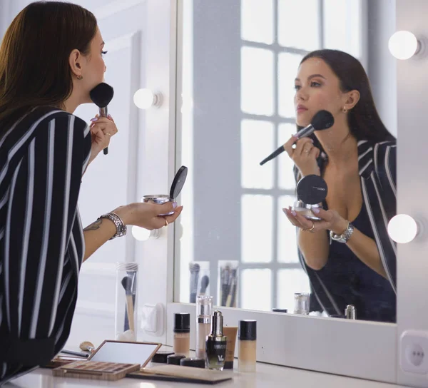 Attractive young woman doing make-up while looking at the mirror and .