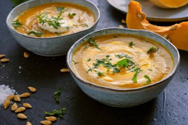 Ev yapımı kabak kremalı çorba maydanozlu ve zeytinyağlı rustik kasenin üstünde çiğ kabak parçaları ve siyah mutfak masasında tohumlar.