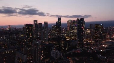 Alacakaranlıktaki Canary Wharf iş merkezindeki gökdelenlerin yükselen görüntüleri. Pembe alacakaranlık gökyüzüne karşı aydınlık binalar. Londra, İngiltere