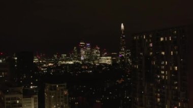 İleri sarmalar gece şehir mahallelerinin üzerinde uçar. Şehir merkezindeki parlayan gökdelenleri gösteriyor. Londra, İngiltere
