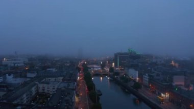 Liffey nehri boyunca şehrin üzerinden geriye doğru uç. Sisli bir akşamda hava görüntüsü. Aydınlatılmış rıhtım. Dublin, İrlanda