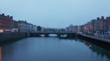 Alacakaranlıkta Liffey nehrinin yukarısında alçak uçuş. Köprüde yürüyen yayalar. Su yüzeyindeki yansımalar. Dublin, İrlanda