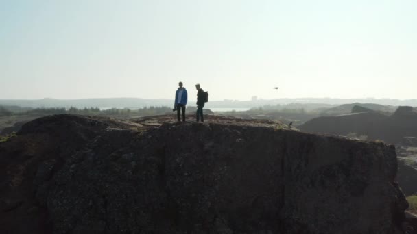 Drönare utsikt över två män som står över klippan ser panorama på Island. Drone utsikt över två backpackers njuter av landskapet i Island efter klättring kulle — Stockvideo