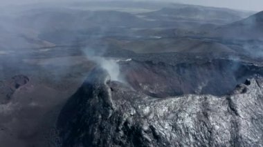Volkan kraterinin yakın çekim görüntüleri. Tepe yamacındaki lav tabakasının metalik parlak yüzeyi. Havalandırmadan beyaz duman yükseliyor. Fagradalsfall volkanı. İzlanda, 2021