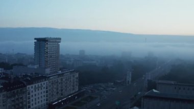 Apartman binaları ve kamu parkı ile kaplı yolun kaydırak ve pan görüntüleri. Şehirde mavi tonlu sisli bir sabah. Varşova, Polonya