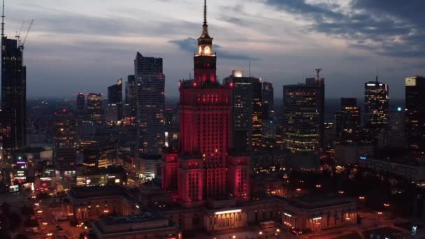 Βραδινό slide και pan πλάνα από κόκκινο φωτισμένο εμβληματικό Παλάτι Πολιτισμού και Επιστημών. Ουρανοξύστες στο κέντρο της πόλης στο βάθος. Βαρσοβία, Πολωνία — Αρχείο Βίντεο