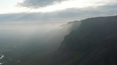 İzlanda 'nın doğal manzarasının hava panoramik görüntüsü. Kara bulutların arasından gelen güneş ışınlarıyla yüksek dağlara doğru uçuyor..