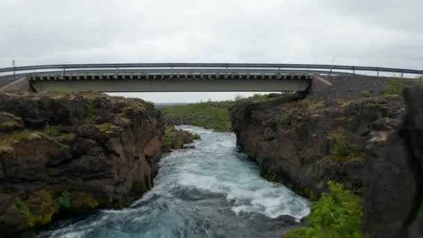 Drone vista aerea che vola sotto il ponte sul fiume che scorre nella campagna rocciosa in Islanda. Occhio di uccello spettacolari altopiani ghiacciati con fiume potentemente scorre — Video Stock