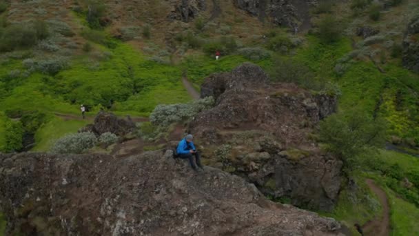 Flygfoto man trekker sitter topp i Island höglandet med hjälp av drönare. Fåglar syn man turist på toppen bergstopp formation i island körning drönare med pad — Stockvideo