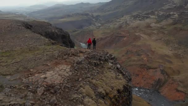 Drohnen betrachten Wanderer, die oben an der Klippe stehen und atemberaubende Ausblicke auf Island genießen. Luftaufnahme zwei Touristen betrachten erstaunliche Landmannalaugar Tal mit Fossa Fluss fließt bemoosten Hochland — Stockvideo