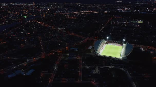 Incrível tiro aéreo de arena de futebol brilhante contrastando com a cidade noturna. Luz brilhante brilhando no campo de jogos de futebol verde. Limerick, Irlanda — Vídeo de Stock