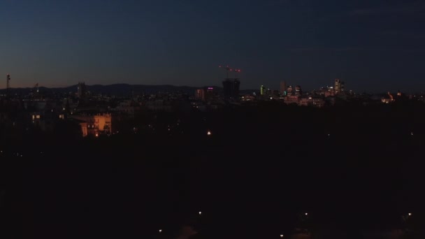 Вечірній політ над деревами в парку. Освітлені будівлі в місті після заходу сонця. Чисте небо в сутінках . — стокове відео