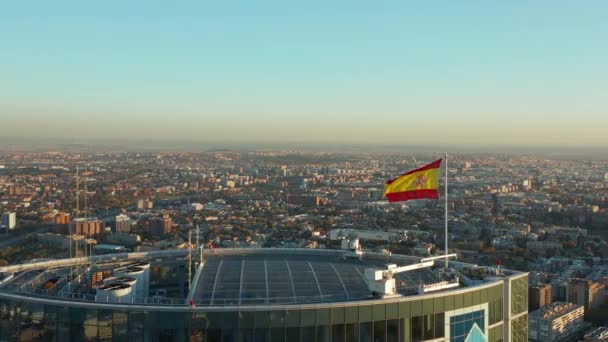 비즈니스 초고층 빌딩의 옥상에서 바람에 흔들며 스페인 국기 주위를 날아다니고 있습니다. 해 가지면서 도시 풍경을 공중에서 들여다본 모습. — 비디오