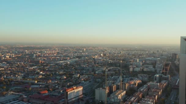 Backwards reveal of top of futuristic high rise office towers. Spanish national flag on pole on rooftop. Aerial view of city at golden hour. — Stock Video