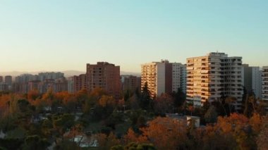 Şehirdeki konutlarda sonbahar öğleden sonra. Parktaki renkli ağaçların üzerinde ileriye doğru uçar. Batan güneşle aydınlatılan apartmanlar.