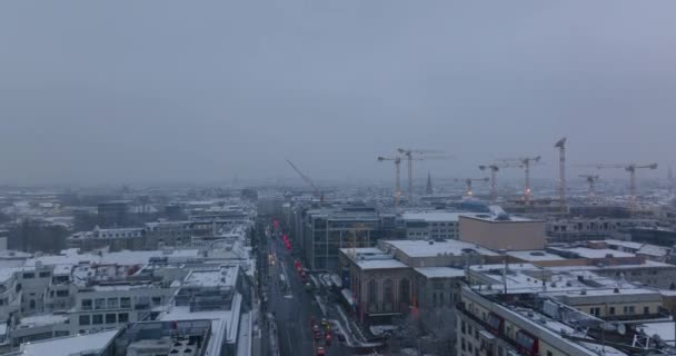 Вид с воздуха на снежные здания в большом городе. Группа башенных кранов на строительной площадке. Машины едут по улице в сумерках. Берлин, Германия — стоковое видео