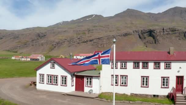 Orbit drone melihat sekitar bendera berkibar dalam angin dari kantor publik di pedesaan Islandia. Pandangan udara Bendera Islandia melambai keluar dari rumah khas di dataran tinggi icelandic — Stok Video