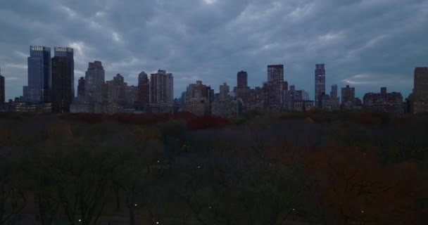 Forwards fly above autumn colour trees in Central park at dusk. Panoramic view of high rise buildings around urban park. Manhattan, New York City, USA — Stock Video