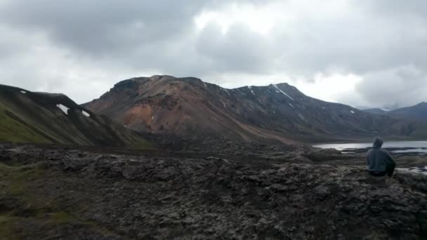Птахи дивляться на людину в зимовій светрі, сидячи на скелі, спостерігаючи за панорамою Ісландських нагір'їв. Чоловік, що сидить на краю скелі, спокійний і спокійний, насолоджуючись панорамою. — стокове відео