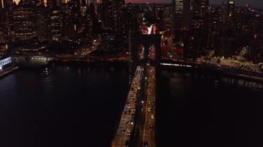Gece Brooklyn Köprüsü 'nün üzerinde uç. Nehir üzerindeki büyük köprüde yavaşça hareket eden araçlar. Rıhtımdaki yüksek binaların ışıklı pencereleri. Manhattan, New York City, ABD
