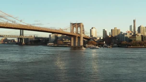Vuelo bajo sobre el agua, crucero flotando en la superficie bajo el puente de Brooklyn. Edificios altos en el fondo. Brooklyn, Nueva York, Estados Unidos — Vídeo de stock