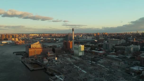 Widok z lotu ptaka na teren przemysłowy nad brzegiem rzeki. Podstacja elektroenergetyczna, magazyn i elektrownia z kominem. Miasto oświetlone przez zachodzące słońce. Brooklyn, Nowy Jork, USA — Wideo stockowe