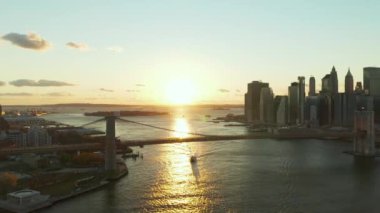 Gün batımına karşı Brooklyn Köprüsü 'nün havadan panoramik görüntüsü. Parlayan su yüzeyinde yüzen bir gemi. Rıhtımdaki gökdelenler. Manhattan, New York City, ABD