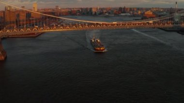 Büyük bir kargo gemisi gün batımında Manhattan Köprüsü 'nün altından geçiyor. Köprü yapısı son güneş ışınlarıyla aydınlandı. Brooklyn, New York City, ABD