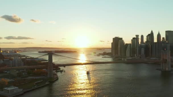 Gün batımına karşı Brooklyn Köprüsü 'nün havadan panoramik görüntüsü. Parlayan su yüzeyinde yüzen bir gemi. Rıhtımdaki gökdelenler. Manhattan, New York City, ABD — Stok video