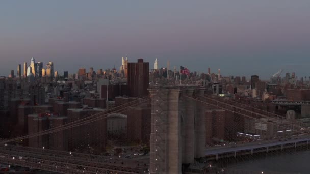 Bandeira americana acenando no vento no topo da torre da ponte. Cityscape com arranha-céus edifícios brilhantes que refletem o céu crepúsculo colorido. Manhattan, Nova Iorque, EUA — Vídeo de Stock