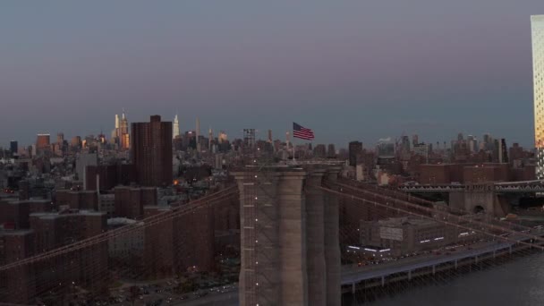 Fly γύρω από την υποστήριξη πύργος της γέφυρας του Μπρούκλιν με ύψωσε αμερικανική σημαία το σούρουπο. Φωτισμένα ψηλά κτίρια σε απόσταση. Μανχάταν, Νέα Υόρκη, ΗΠΑ — Αρχείο Βίντεο