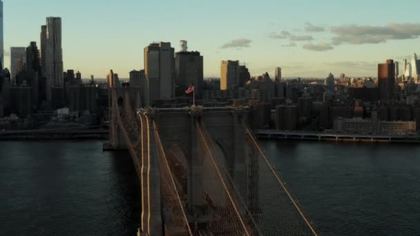 Флаг США, размахивающий на Бруклинском мосту на закате. Удивительный вид на современные высотные здания в центре города. Манхэттен, Нью-Йорк, США — стоковое видео