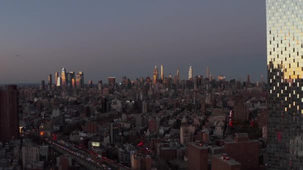 Fly above city at dusk, tall illuminated skyscrapers in distance. Sliding reveal of glossy high rise building reflecting colourful sunset sky. Manhattan, New York City, USA — Vídeo de Stock