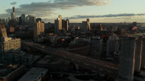 日没の時間に都市の空中ビュー。アメリカ国旗が掲揚された古い橋の上を走る複々線の道路が混雑している。米国ニューヨーク市ブルックリン区 — ストック動画