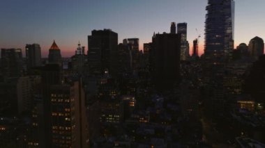 Alacakaranlıkta şehrin üzerinde uç. Işıklı pencereli yüksek binalar. Renkli gökyüzüne karşı siluetler. Manhattan, New York City, ABD