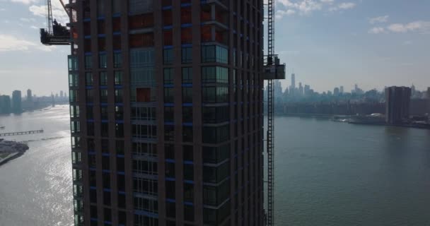 Pull back shot of construction of new modern tall skyscraper on riverbank. Construction lift moving down around wall. Downtown skyscrapers in distance. New York City, USA — Stock Video