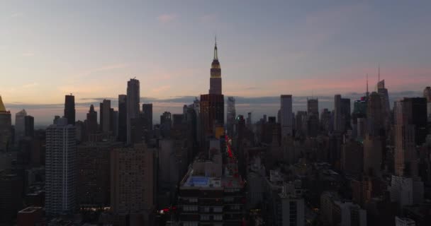 夕方の街の上を飛ぶ。カラフルな夕暮れの空に対する高層ビルの上。夕暮れ時にエンパイアステートビルのライトアップされた尖塔。米国ニューヨーク市マンハッタン — ストック動画