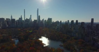 Sonbahar Central Park 'ın üzerinde renkli ağaçlar ve güneşi yansıtan su yüzeyiyle uçun. Arka planda modern şehir merkezi ofis kuleleri var. Manhattan, New York City, ABD