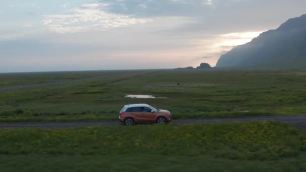 Tracking van SUV auto rijden op onverharde weg op het platteland bij zonsopgang of zonsondergang. Groen vlak grasland omgeven door rotsachtige bergkam. IJsland — Stockvideo
