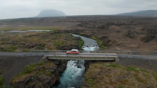 Ziehen Sie Filmaufnahmen von einer schmalen Straßenbrücke über einen Wildwasserbach und einem fahrenden Auto zurück. Eine flache Landschaft. Island — Stockvideo